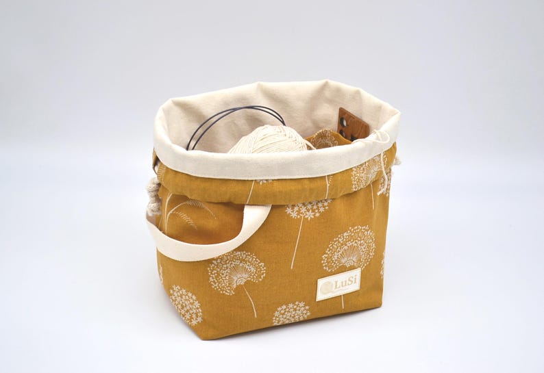 May include: Mustard yellow fabric tote bag with a white dandelion pattern. The bag has a cream-coloured interior and handles. Inside the bag are knitting supplies, including yarn and needles. The label reads "LuSi".