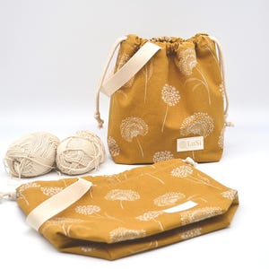 May include: Mustard yellow drawstring bags with a white dandelion pattern. The bags have cream-coloured straps and drawstrings. Two balls of yarn are next to the bags. The bags have the text "LuSi" on a white label.