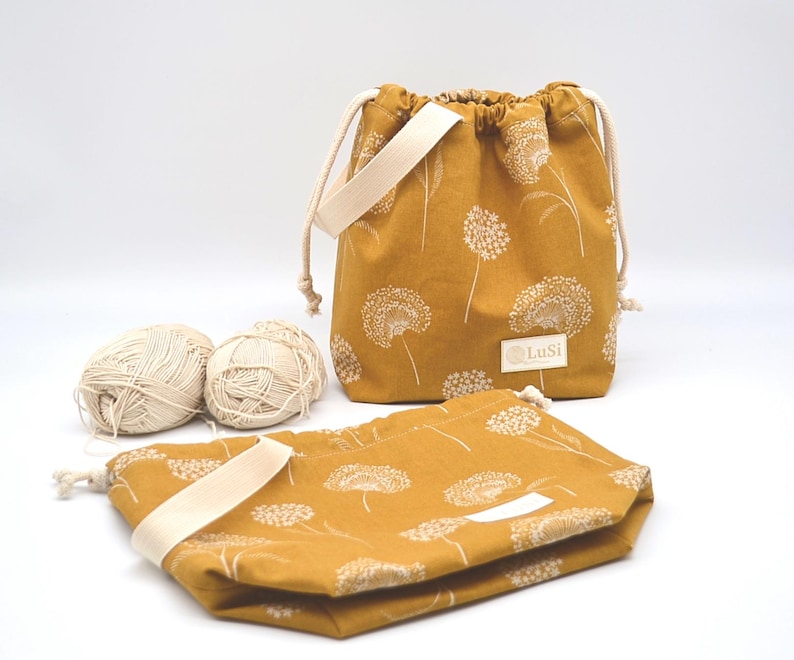 May include: Mustard yellow drawstring bags with a white dandelion pattern. The bags have cream-coloured straps and drawstrings. Two balls of yarn are next to the bags. The bags have the text "LuSi" on a white label.