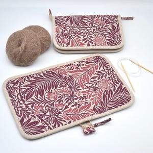 Puede incluir: Dos estuches rectangulares para agujas de tejer con un estampado floral burdeos y rosa, ribete crema y cierre de cremallera. Tambi&eacute;n se ven dos ovillos de lana marr&oacute;n claro y un par de agujas de tejer. Para suministros de tejido.