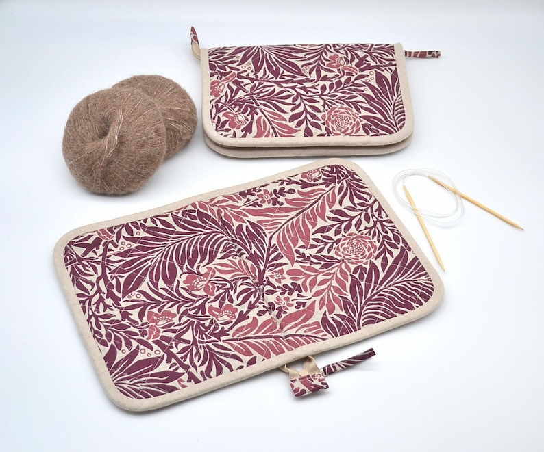 Puede incluir: Dos estuches rectangulares para agujas de tejer con un estampado floral burdeos y rosa, ribete crema y cierre de cremallera. Tambi&eacute;n se ven dos ovillos de lana marr&oacute;n claro y un par de agujas de tejer. Para suministros de tejido.