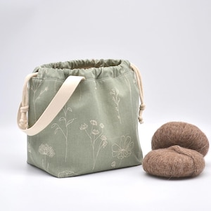 May include: A sage green drawstring bag with a cream-colored strap and floral pattern. Two brown yarn balls sit next to the bag. The bag is designed for carrying knitting or crochet projects.