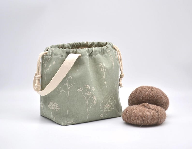 May include: A sage green drawstring bag with a cream-colored strap and floral pattern. Two brown yarn balls sit next to the bag. The bag is designed for carrying knitting or crochet projects.