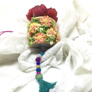 May include: A wooden spool decorated with colourful fabric flowers and green leaves, with a beaded tassel hanging from the bottom. The spool is sitting on a white lace fabric.