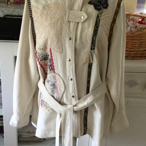 May include: Cream-coloured jacket with decorative patches, lace, and a butterfly accent. Features a mandarin collar, button closure, and a matching belt. The jacket has decorative trim along the sleeves and front.