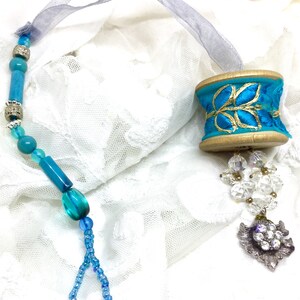 May include: Teal blue and gold ribbon spool bookmark with a crystal and rhinestone charm. The bookmark is strung with teal blue beads and silver spacers.