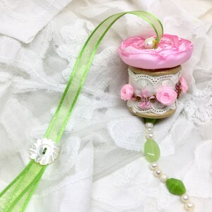 May include: A pink and white spool with a green ribbon and a pearl and green bead necklace. The spool is decorated with lace and pink flowers.