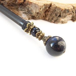 May include: A dark blue hair stick with a large, round, sparkling blue stone at the end. The stick has ornate bronze-colored metal accents and a dark wooden shaft. The background is white with a piece of wood.