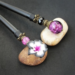 Puede incluir: Dos palos de pelo de madera negra con detalles decorativos. Un palo tiene un encanto de flor rosa y negro y una cuenta de vidrio rosa. El otro palo tiene una cuenta de vidrio negro y una cuenta de vidrio morado.