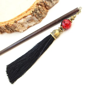 May include: A dark brown wooden hair stick with a black tassel and a red glass bead with gold accents.