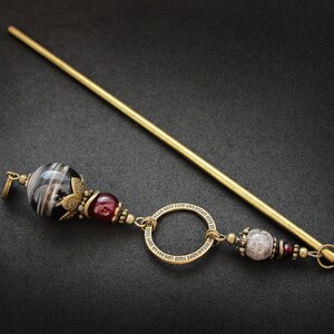 May include: A gold hair stick with a black and white agate bead, a red bead, a circle charm with an inscription, and a white and gray bead.