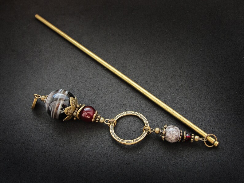 May include: A gold hair stick with a black and white agate bead, a red bead, a circle charm with an inscription, and a white and gray bead.
