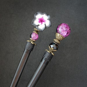 Puede incluir: Dos palos de pelo negros con tapas decorativas. Un palo tiene un dise&ntilde;o de flor rosa y blanca, el otro tiene una esfera de vidrio rosa con detalles dorados.