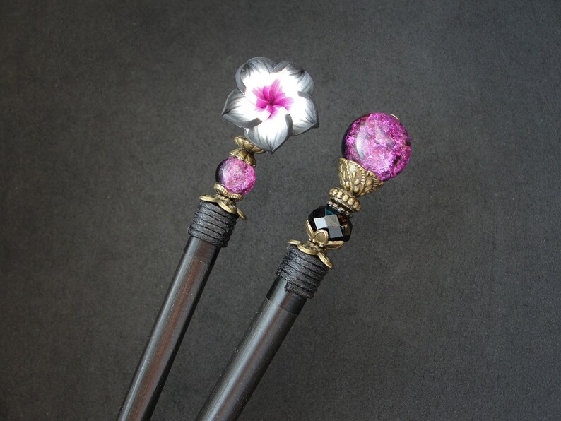 Puede incluir: Dos palos de pelo negros con tapas decorativas. Un palo tiene un dise&ntilde;o de flor rosa y blanca, el otro tiene una esfera de vidrio rosa con detalles dorados.