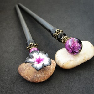 Puede incluir: Dos palos de pelo negros con extremos decorativos. Un palo tiene una flor rosa con detalles blancos y el otro tiene una cuenta de vidrio p&uacute;rpura con detalles negros.