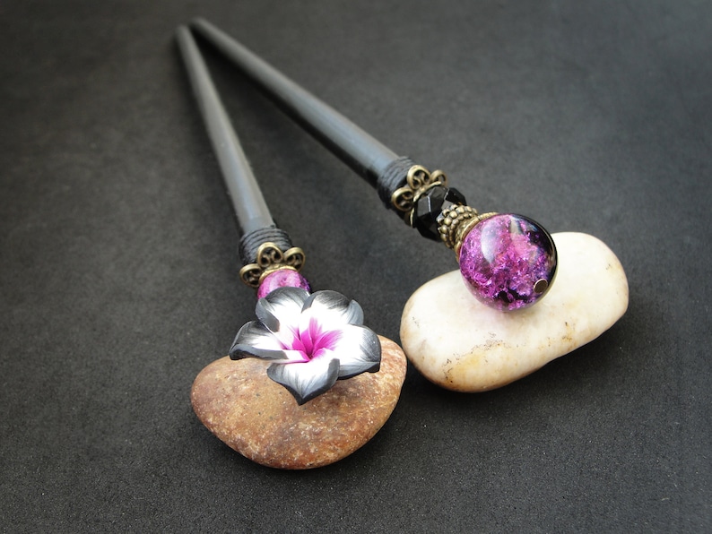 Puede incluir: Dos palos de pelo negros con extremos decorativos. Un palo tiene una flor rosa con detalles blancos y el otro tiene una cuenta de vidrio p&uacute;rpura con detalles negros.