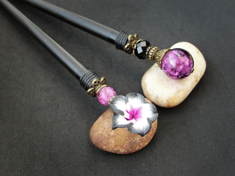 Puede incluir: Dos palos de pelo de madera negra con detalles decorativos. Un palo tiene un encanto de flor rosa y negro y una cuenta de vidrio rosa. El otro palo tiene una cuenta de vidrio negro y una cuenta de vidrio morado.