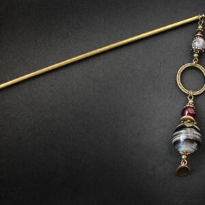 May include: A gold hair stick with a decorative pendant. The pendant features a black and white agate bead, a maroon bead, and a gold ring.