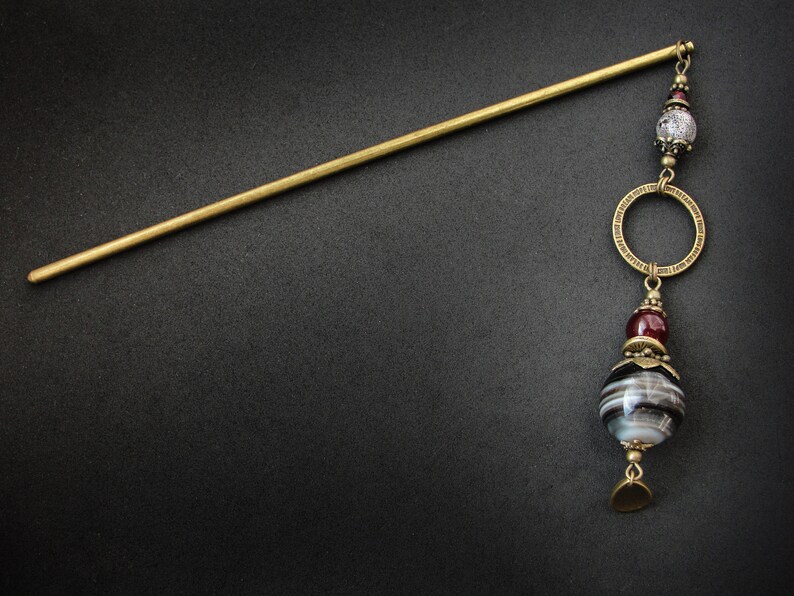 May include: A gold hair stick with a decorative pendant. The pendant features a black and white agate bead, a maroon bead, and a gold ring.