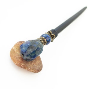 May include: A dark wooden hair stick with a blue gemstone and bronze accents. The stick is resting on a brown stone.