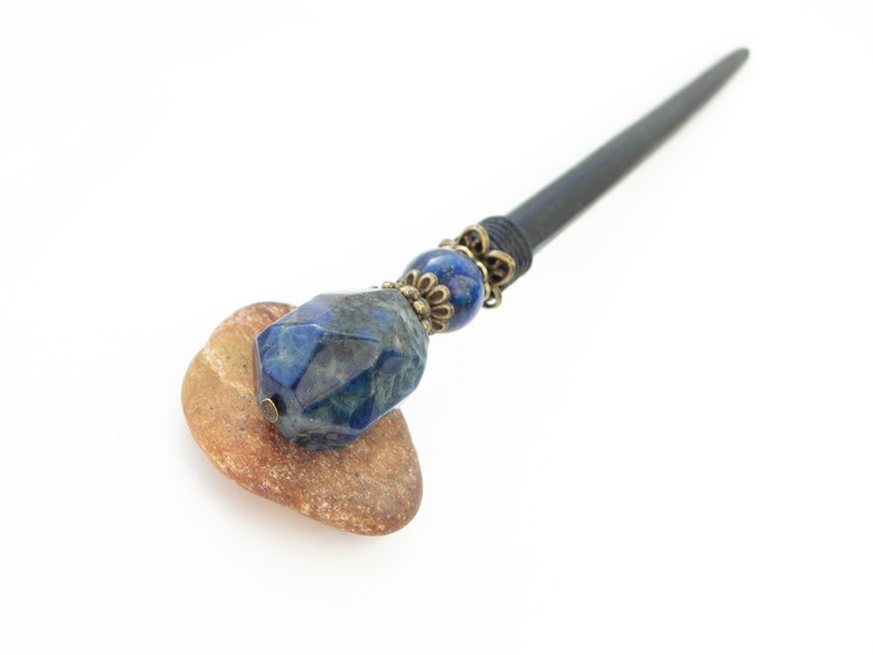 May include: A dark wooden hair stick with a blue gemstone and bronze accents. The stick is resting on a brown stone.