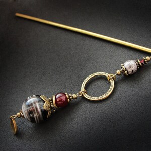 May include: A gold hair stick with a decorative bead and a ring. The bead is white with a textured surface and the ring is round with a detailed design. The stick is about 10 inches long.