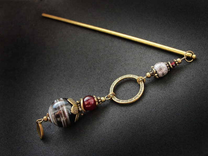May include: A gold hair stick with a decorative bead and a ring. The bead is white with a textured surface and the ring is round with a detailed design. The stick is about 10 inches long.