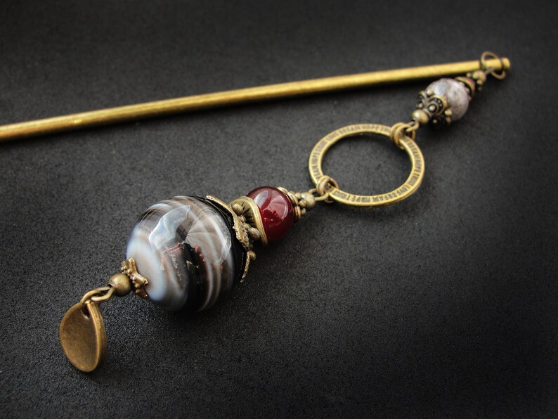 May include: A gold hair stick with a decorative circle, a red and white agate bead, and a smaller white agate bead with a brown and white swirl pattern.