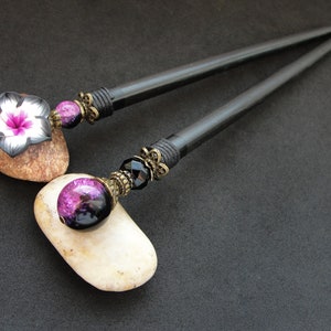 Puede incluir: Dos palos de pelo de madera negra con detalles decorativos. Un palo tiene una cuenta de vidrio p&uacute;rpura y negra y el otro tiene una flor negra y blanca.