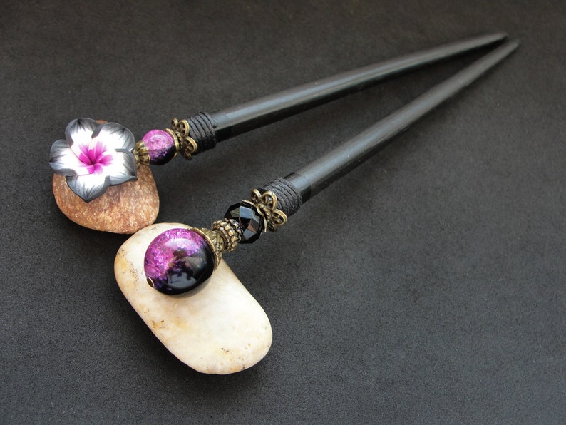 Puede incluir: Dos palos de pelo de madera negra con detalles decorativos. Un palo tiene una cuenta de vidrio p&uacute;rpura y negra y el otro tiene una flor negra y blanca.
