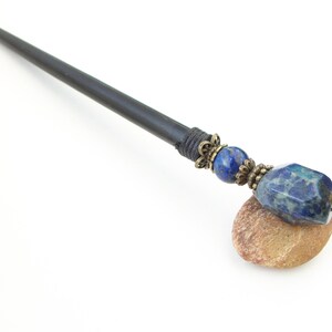 May include: A black wooden hair stick with a blue and gold gemstone accent. The stick is resting on a small brown rock.