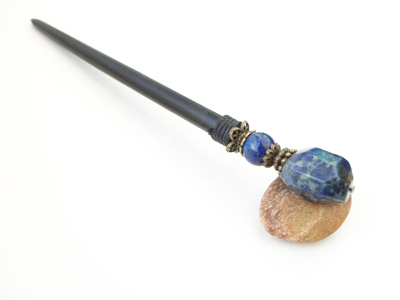 May include: A black wooden hair stick with a blue and gold gemstone accent. The stick is resting on a small brown rock.