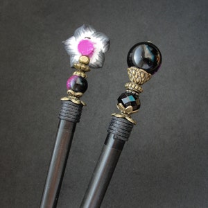 Puede incluir: Dos palos de pelo de madera negra con tapas decorativas. Un palo tiene una flor negra y blanca con un centro rosa. El otro palo tiene una esfera de vidrio negro con un dise&ntilde;o de metal dorado.