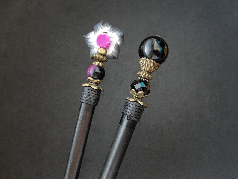 Puede incluir: Dos palos de pelo de madera negra con tapas decorativas. Un palo tiene una flor negra y blanca con un centro rosa. El otro palo tiene una esfera de vidrio negro con un dise&ntilde;o de metal dorado.