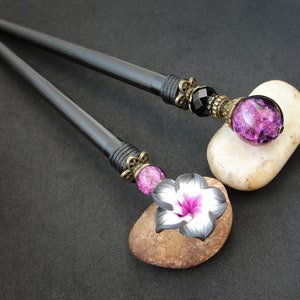 Puede incluir: Dos palos de pelo de madera negra con detalles decorativos. Un palo tiene un encanto de flor rosa y negro, el otro tiene una cuenta de vidrio negro y morado.