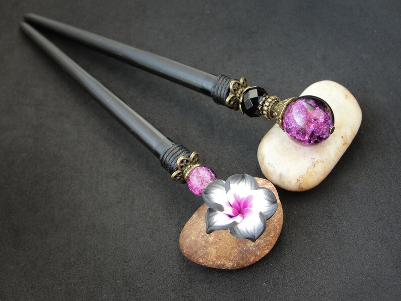 Puede incluir: Dos palos de pelo de madera negra con detalles decorativos. Un palo tiene un encanto de flor rosa y negro, el otro tiene una cuenta de vidrio negro y morado.
