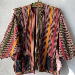 May include: A colorful, open-front jacket with vertical stripes in red, orange, green, and purple. The jacket has two black pockets and a mandarin collar. The sleeves are three-quarter length. The jacket is hanging on a decorative wooden hanger.