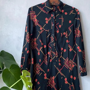 Può includere: Abito nero a maniche lunghe con colletto e chiusura a bottoni. L'abito presenta un motivo geometrico rosso e oro. È appeso a una gruccia di legno. L'abito ha un corpetto aderente e una gonna leggermente svasata.