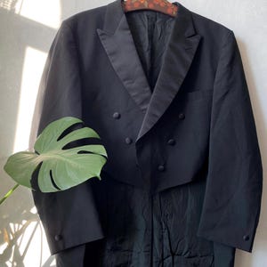 May include: A black tuxedo jacket with a double-breasted front and a notched lapel. The jacket has six buttons and a long tail. The jacket is hanging on a wooden hanger. A large green leaf is in the foreground.