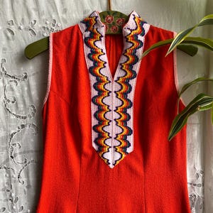 May include: A sleeveless, orange dress with a V-neck. The neckline is trimmed with a white, scalloped design featuring a rainbow of colours. The dress is hanging on a green hanger.