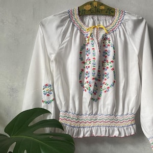 Pode incluir: Blusa branca de manga comprida com bordados florais coloridos. A blusa apresenta cintura e decote franzidos, com um laço amarelo na gola. As mangas têm punhos bordados. O bordado inclui fio vermelho, azul, amarelo e verde.