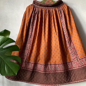 May include: An orange skirt with a paisley and teardrop pattern. The skirt has a brown quilted hem and a brown waistband. The skirt is made of a lightweight fabric and has a vintage appearance.