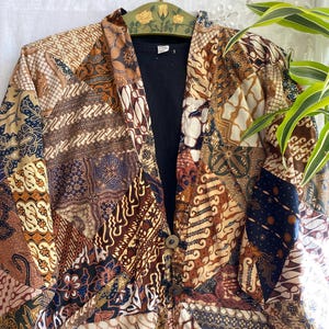 Puede incluir: Chaqueta patchwork con una variedad de patrones batik marrones, beige y azul marino. La chaqueta tiene cuello y tres botones de madera. Se ve una camiseta negra debajo.