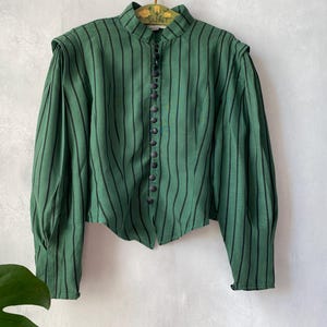 May include: Green and black striped blouse with a high collar and button-down front. The long-sleeved top features shoulder detailing and a fitted silhouette. The fabric has vertical black stripes on a green background. The blouse is hanging on a vintage hanger.