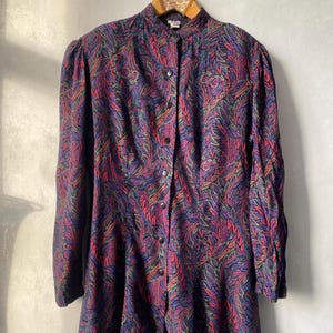 Puede incluir: Un vestido vintage con botones y un vibrante estampado de cachemira en tonos morado, rojo, verde y azul. El vestido presenta mangas largas, cuello alto y una silueta entallada. El vestido está colgado en una percha de madera.