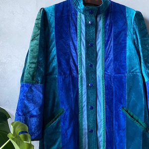 Può includere: Giacca vintage a blocchi di colore blu e verde. Colletto alto, chiusura con bottoni e tasche applicate. Il tessuto sembra velluto, con strisce verticali di diverse larghezze. Un look retrò anni '70.
