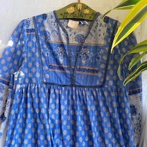 Puede incluir: Vestido azul vintage con cuello en V y mangas largas. El vestido presenta un estampado floral y a rayas en tonos azules, beige y marrón. El vestido está colgado de una percha de madera con detalles florales.