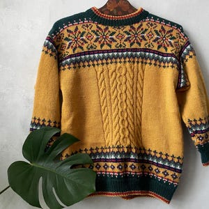 Peut inclure: Pull-over jaune moutarde avec un motif en tricot torsadé sur le devant. Le pull a un col rond et est orné d'un motif géométrique vert, rouge, blanc et jaune. Les poignets et l'ourlet sont verts avec un motif blanc et rouge.