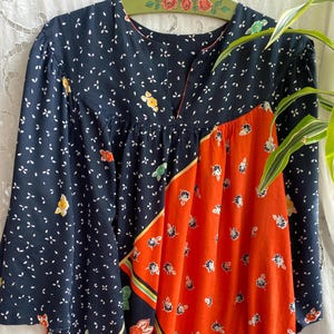 Può includere: Un abito vintage blu navy con scollo a V e maniche lunghe. L'abito presenta un motivo floreale con accenti bianchi, gialli e arancioni. Una sezione arancione presenta una stampa floreale e un bordo verde, giallo e nero.