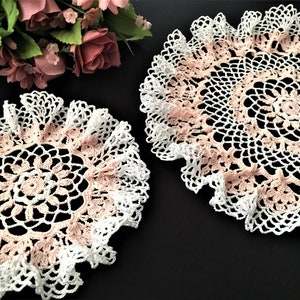 Op de afbeelding: Twee delicate gehaakte doilies in wit en roze met een gegolfde rand. De doilies zijn gemaakt met een fijn garen en hebben een delicaat bloemenpatroon.